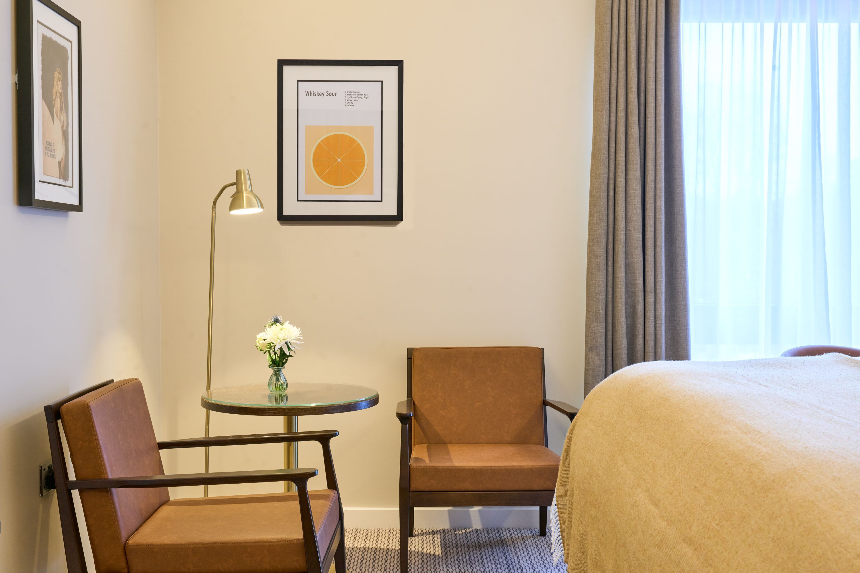Een hotelkamer met een bed, twee stoelen, een tafeltje met een bloevas, een lijst met een afbeelding van een sinaasappel en een lamp.