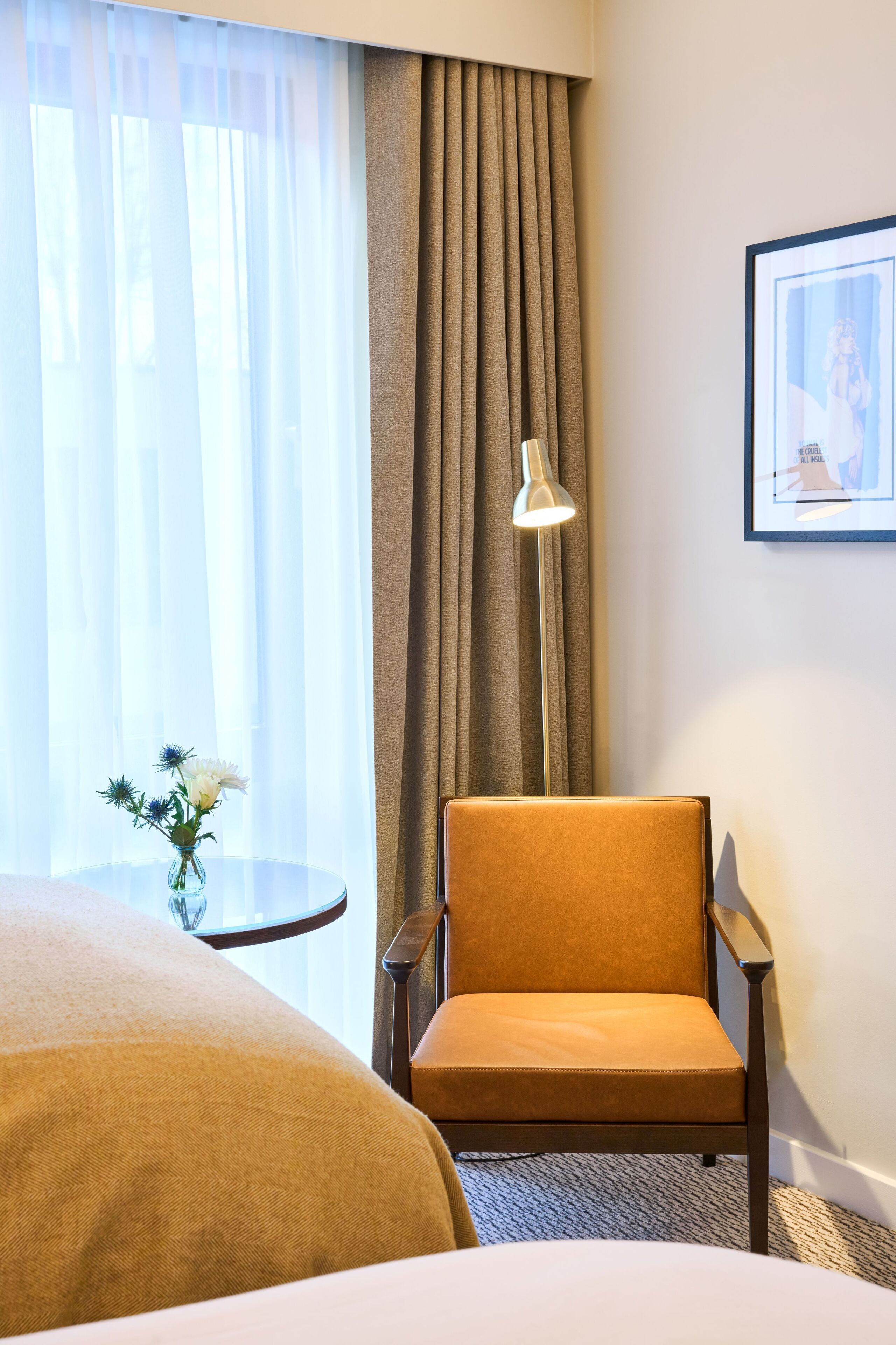 Habitació d'hotel amb un llit, una cadira, una taula petita amb un vas de flors, una làmpada i un quadre al mur.