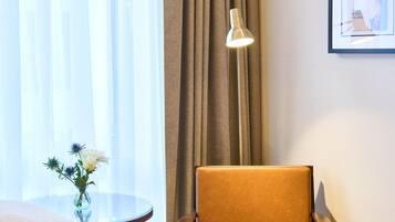 Habitación de hotel con cama, una silla, una mesita con un florero y una lámpara, además de un cuadro en la pared.