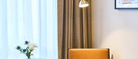 A hotel room with a bed, a chair, a small table with a vase of flowers, a lamp, and a framed picture on the wall.