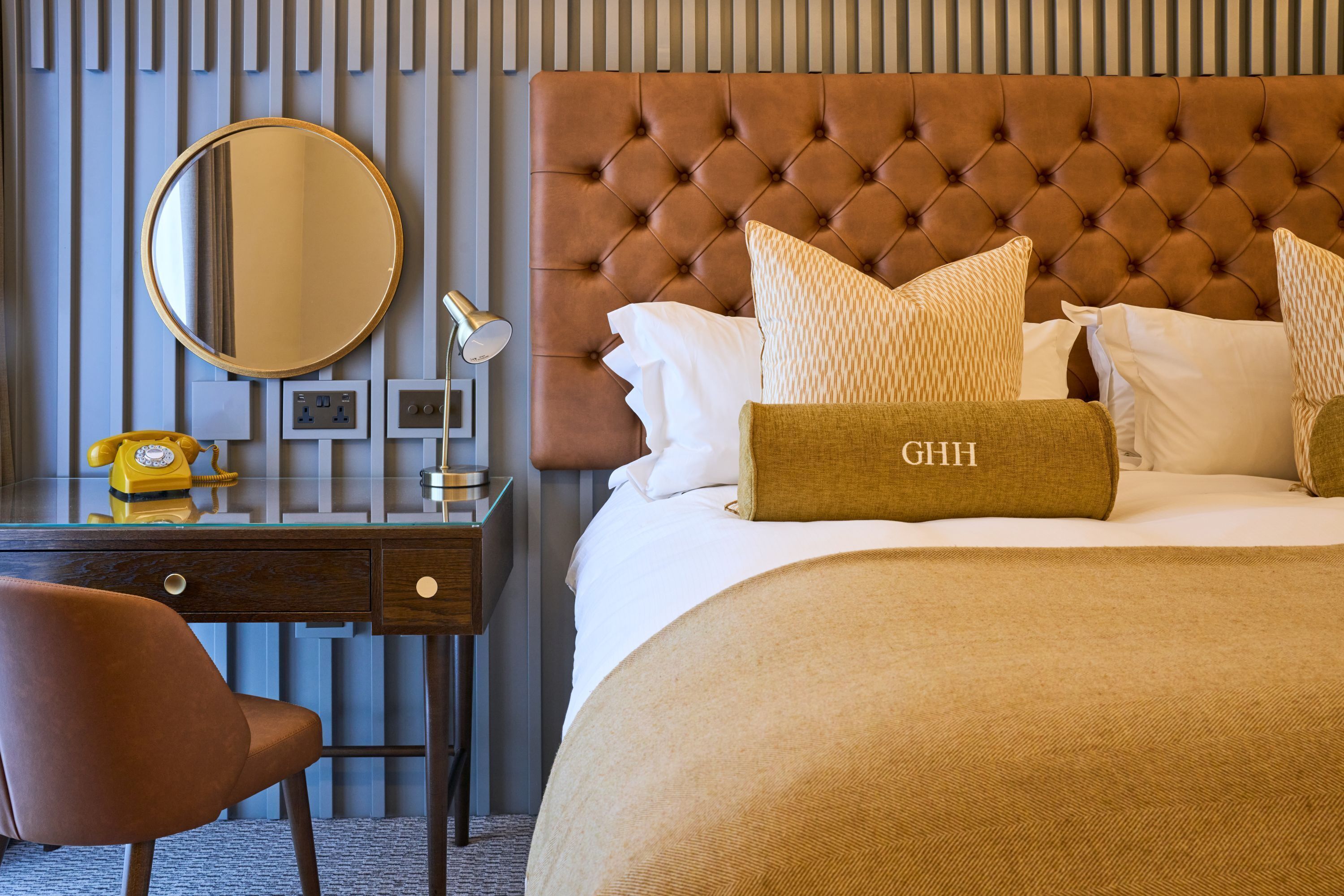A hotel room with a tufted headboard, a round mirror, a desk with a telephone, and a bed with a beige blanket and pillows.