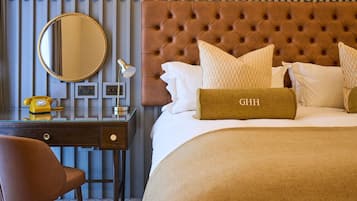 Une chambre d’hôtel avec une tête de lit capitonnée, un miroir rond, un bureau avec un téléphone et un lit recouvert d’une couverture beige et agrémenté d’oreillers.