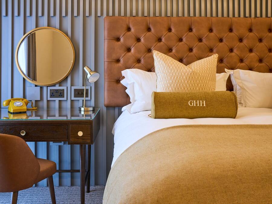 Une chambre d’hôtel avec une tête de lit capitonnée, un miroir rond, un bureau avec un téléphone et un lit recouvert d’une couverture beige et agrémenté d’oreillers.