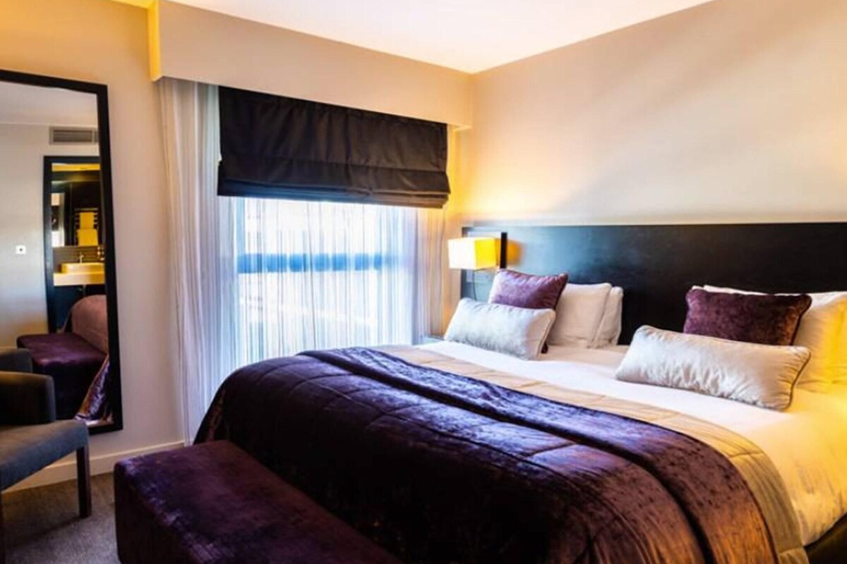 Une chambre d’hôtel avec un grand lit, une table de chevet, une chaise et une fenêtre avec des rideaux.