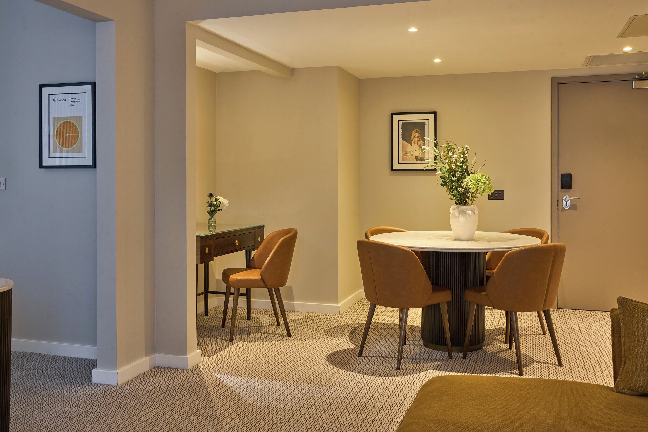 A hotel room with a round dining table, four chairs, a vase with flowers, a framed picture on the wall, and a door with a lock.