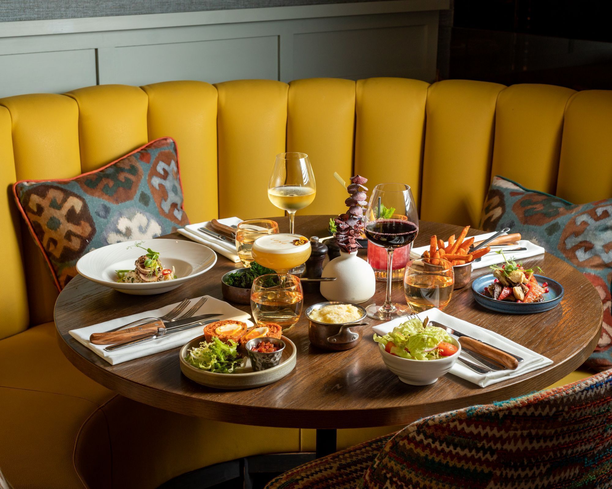 A round dining table set with various dishes, glasses of wine, and cutlery, surrounded by a yellow upholstered sofa with patterned cushions.