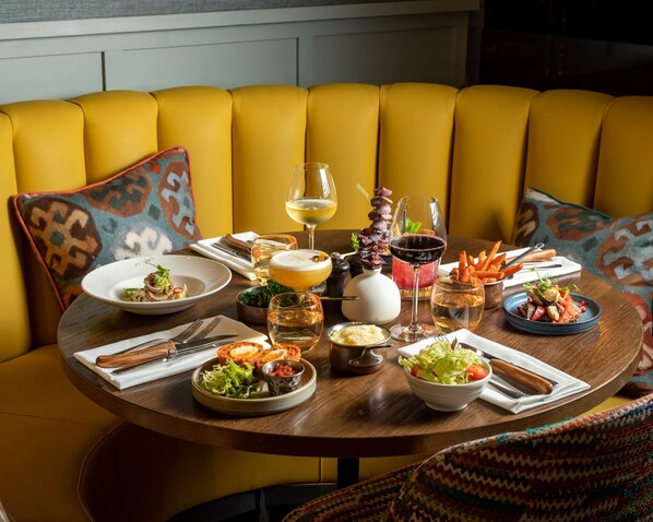 A round dining table set with various dishes, glasses of wine, and cutlery, surrounded by a yellow upholstered sofa with patterned cushions. - Harbour Hotel Guildford (Guildford)