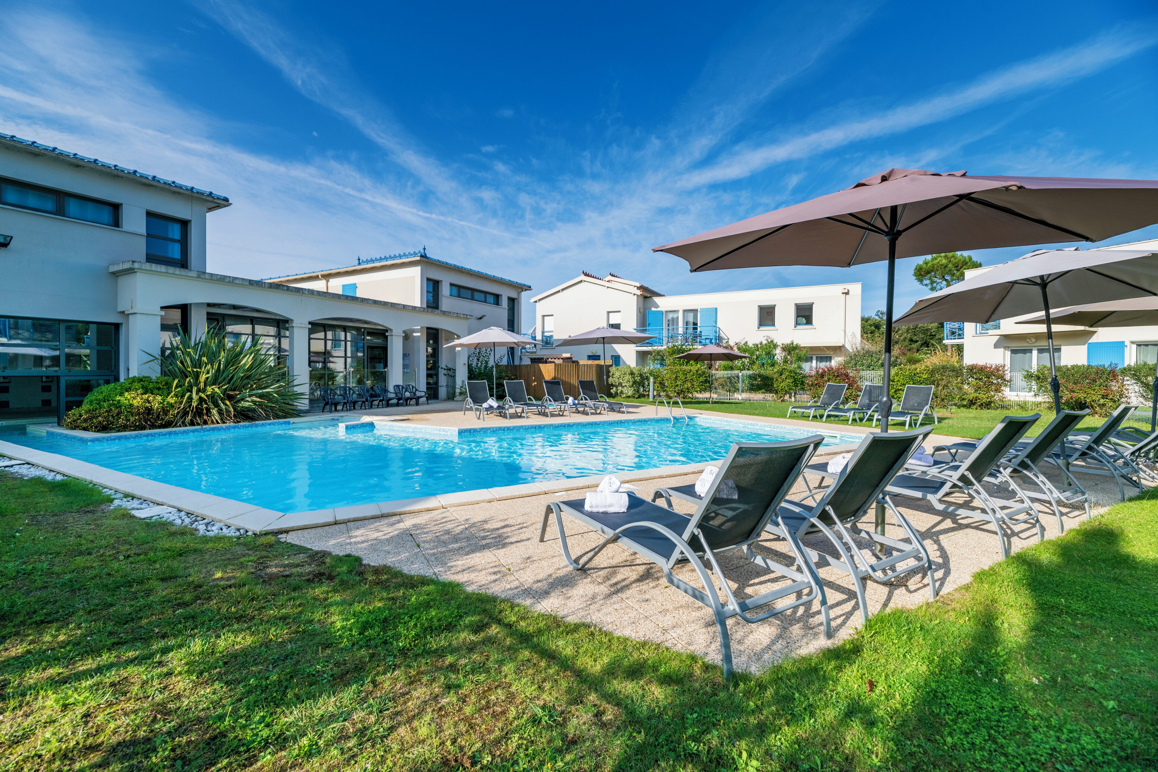 A swimming pool with lounge chairs and umbrellas, surrounded by modern buildings and greenery.