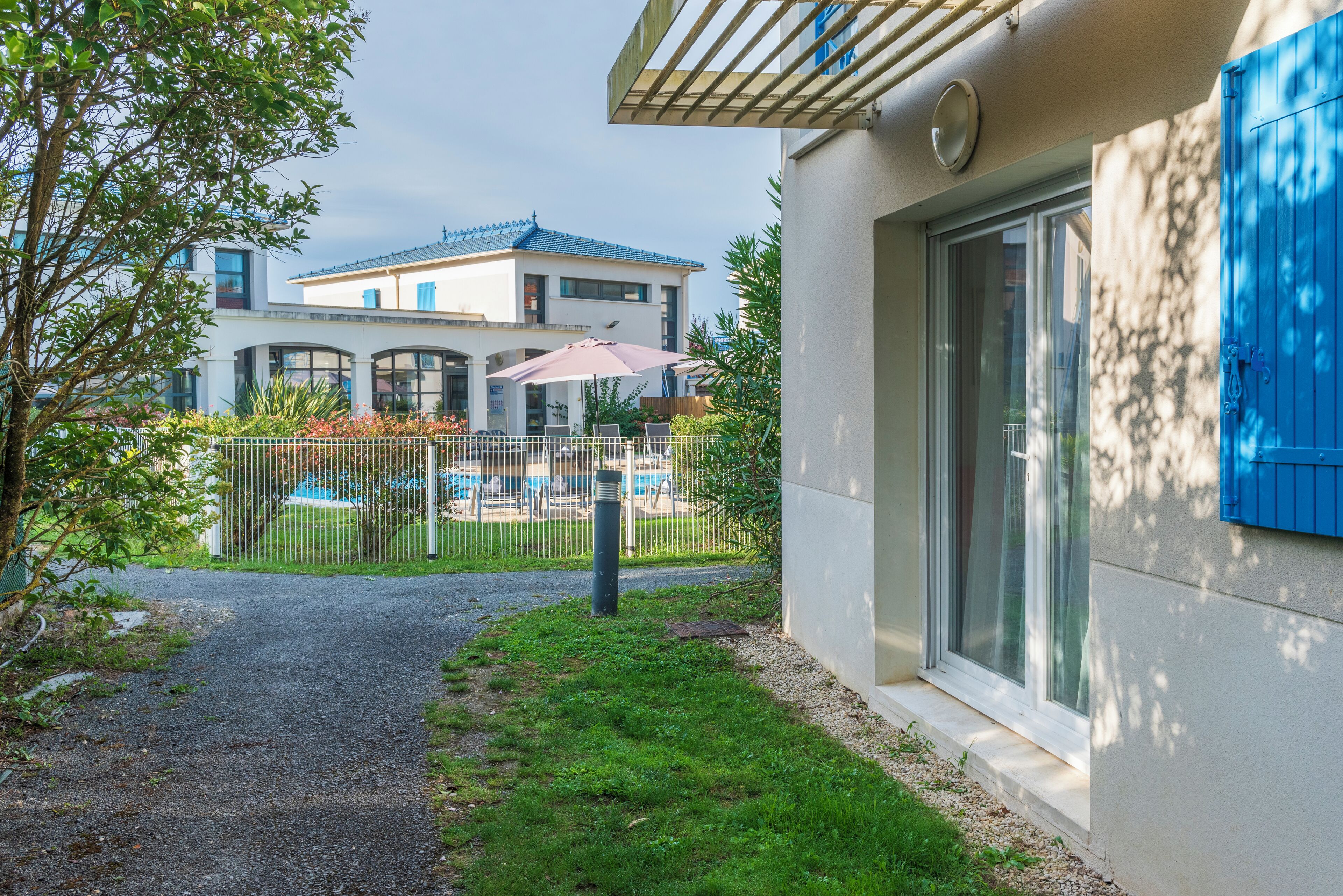 A pathway leading to a modern house with a blue door, a visible swimming pool, and a fenced garden area.