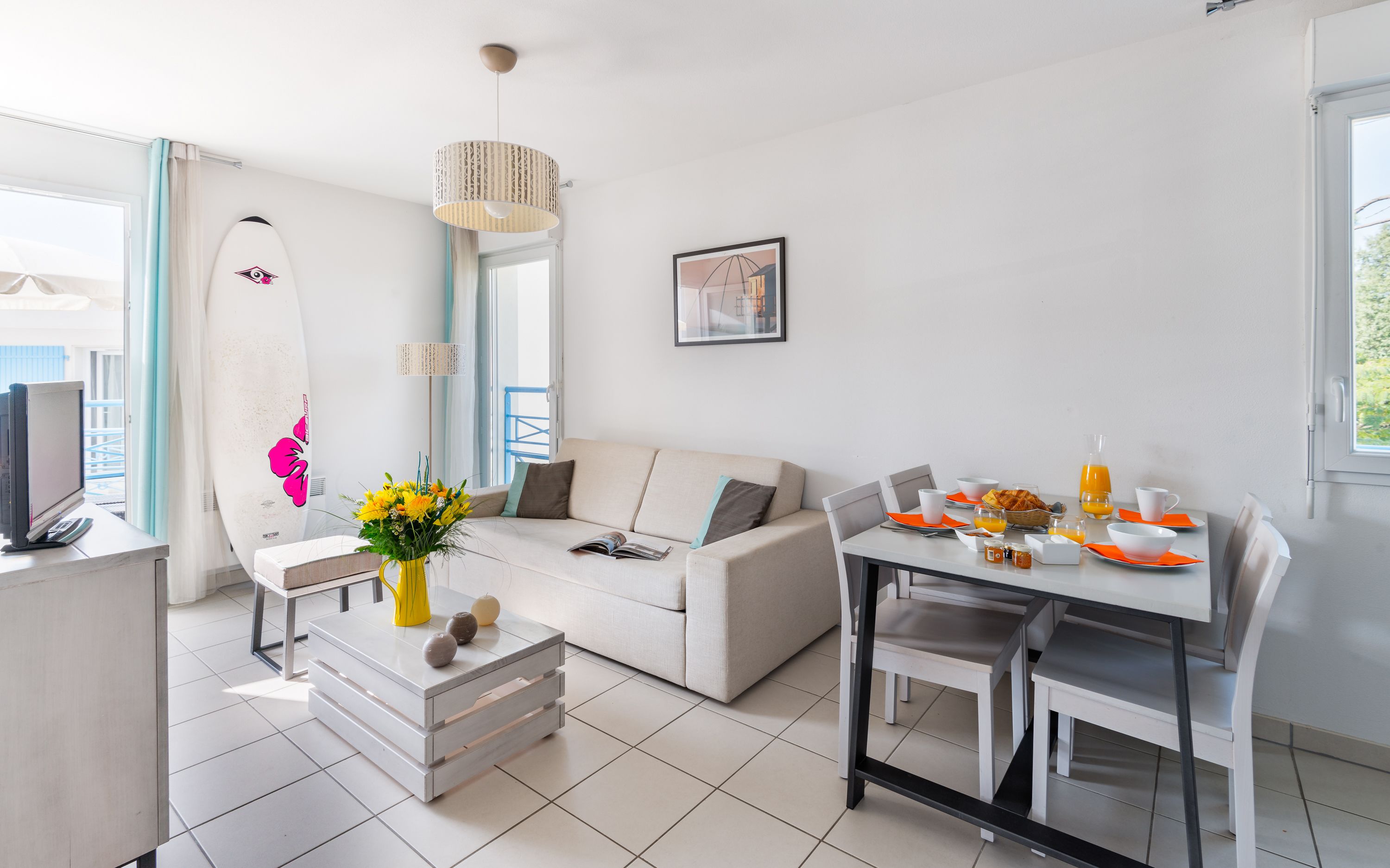 Ein modernes Wohnzimmer mit einem weißen Sofa, einem Esstisch mit orangefarbenen Untersetzern, einem Couchtisch mit Dekoration und einem großen Fenster mit Vorhängen.
