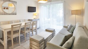 Una sala de estar moderna con un sofá, una mesa de comedor con sillas, un televisor montado en la pared y un ventanal grande.