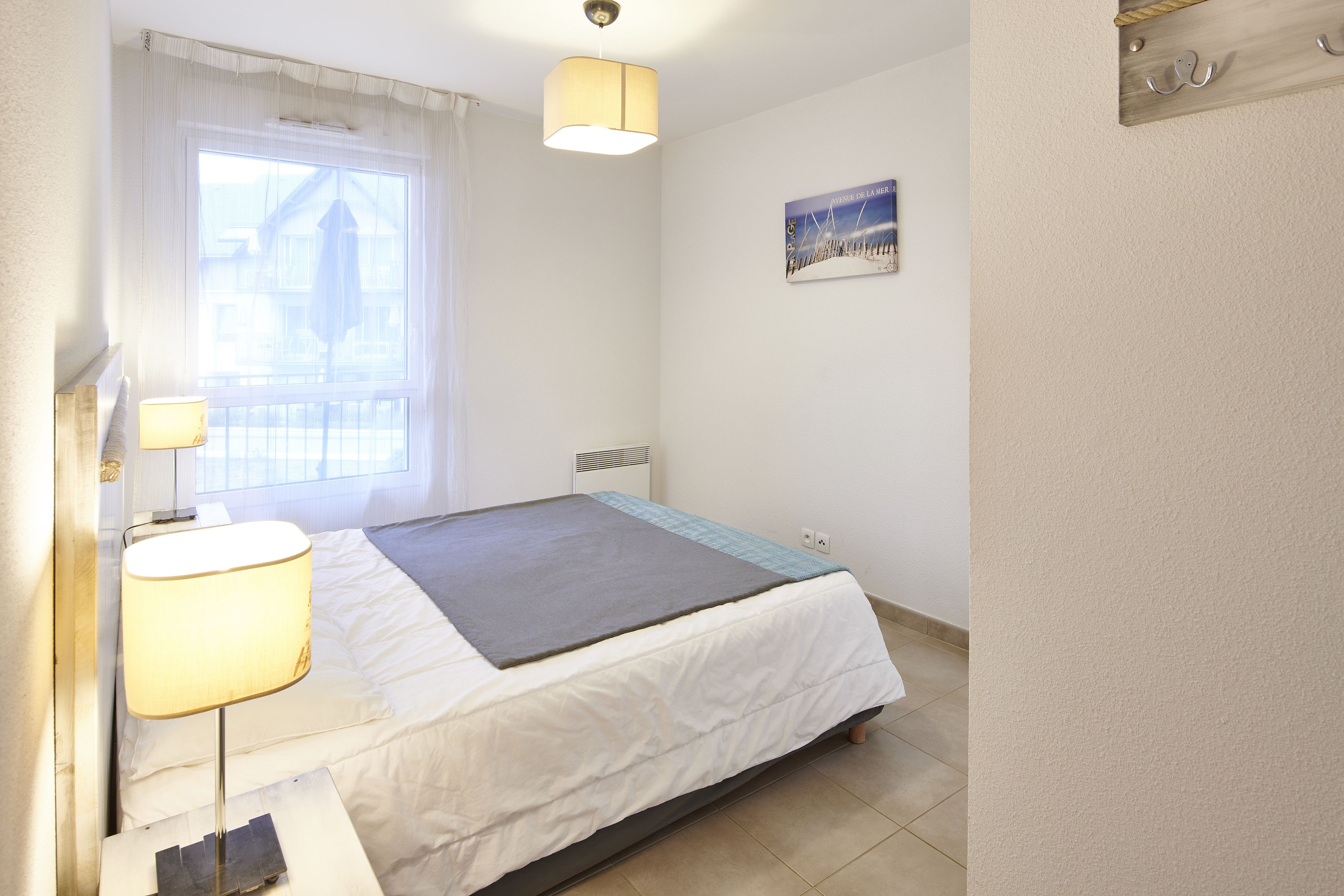 A neatly arranged bedroom with a bed, bedside lamps, a window with curtains, a wall-mounted hookboard, and a small picture.