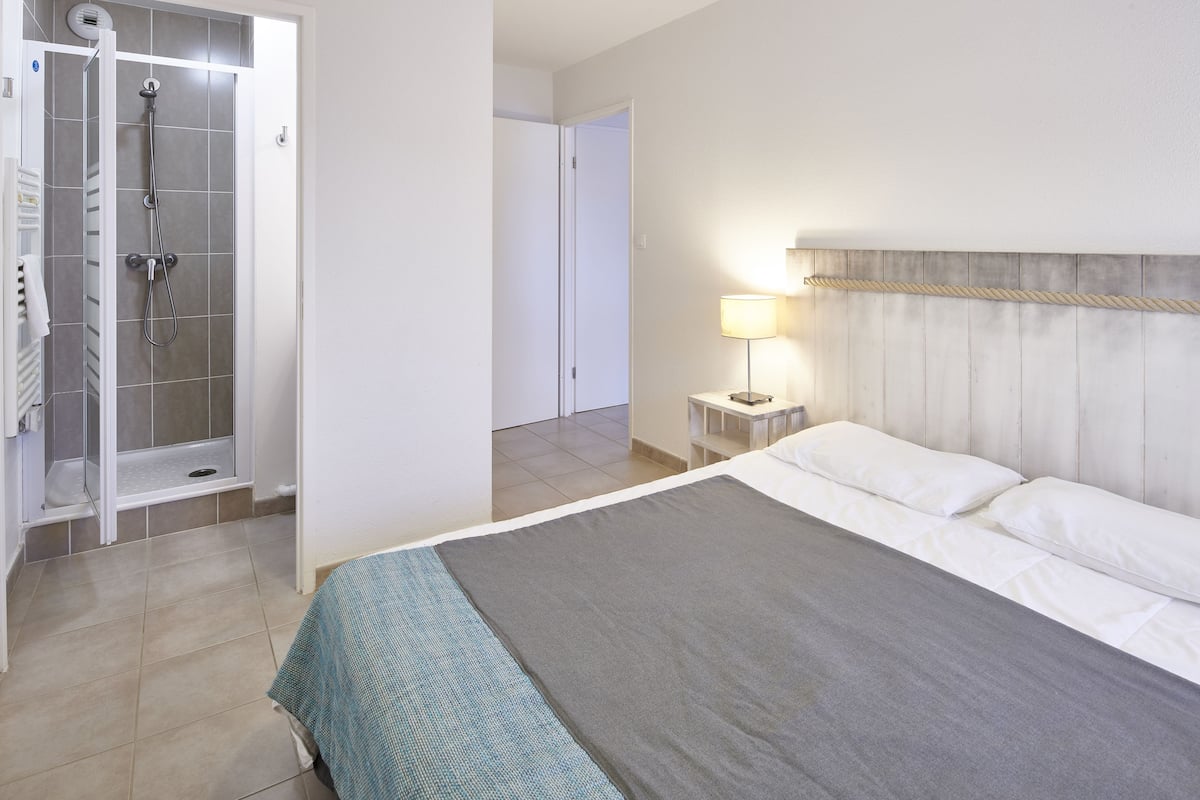 A hotel room with a bed, a shower, a bedside table with a lamp, and a view of the hallway.