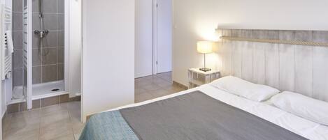 Una habitación de hotel con cama, ducha, mesita de noche con lámpara y vista al pasillo.