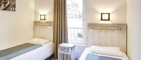 A hotel room with two single beds, a small table, and a window with curtains.