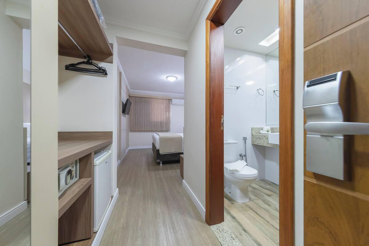 A narrow hallway leading to a bathroom with a toilet, sink, and shower. A bed is visible in the background.