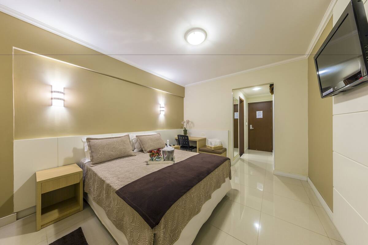A hotel room with a bed, bedside table, lamp, TV, and a door leading to another room.