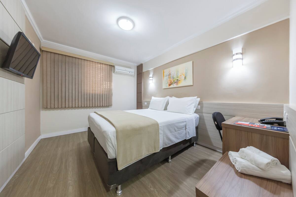 A hotel room with a bed, a desk with a telephone, a chair, a flat-screen TV mounted on the wall, and a painting on the wall.