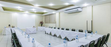 Un salón de conferencias con largas mesas, sillas y botellas de agua.