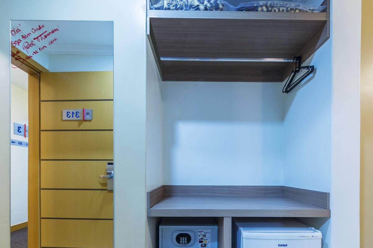 A compact room with a wooden door marked with a number, a small refrigerator, and a shelf with a plastic bag.