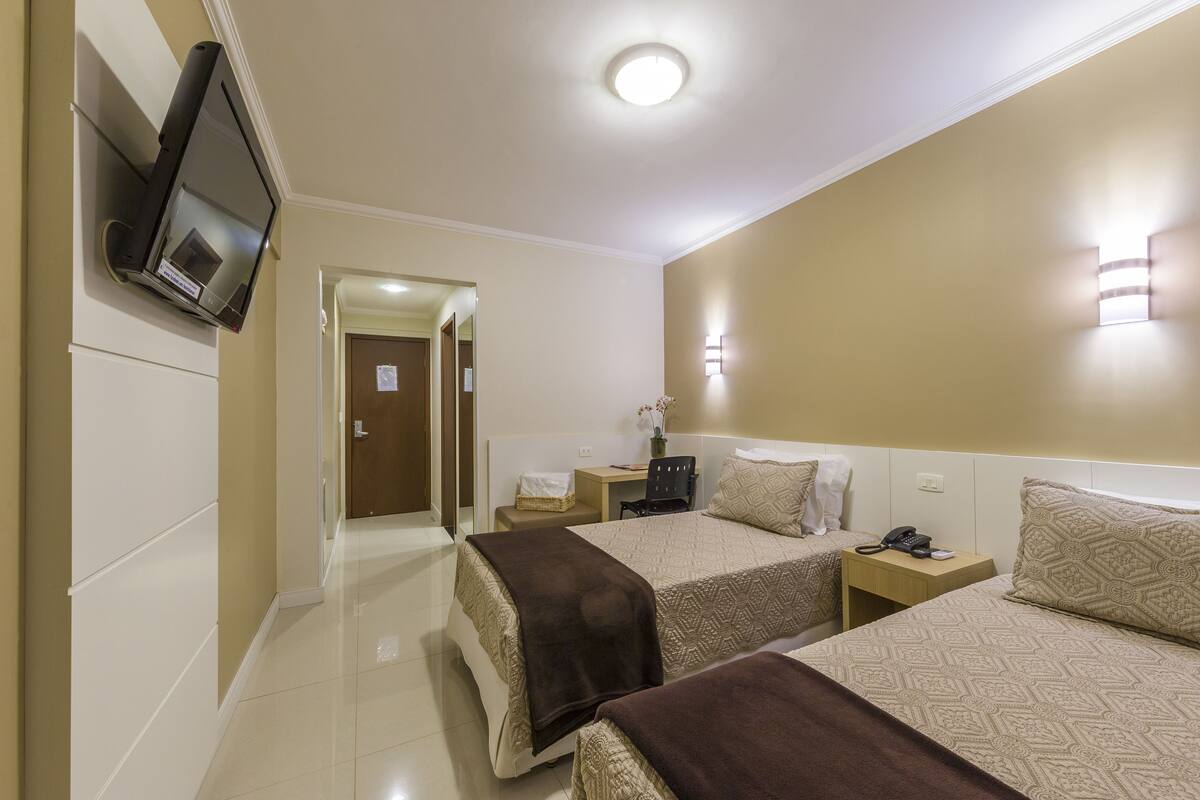 A hotel room with two beds, a TV mounted on the wall, a bedside table with a lamp, and a door leading to another room.
