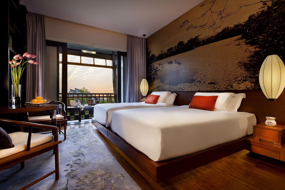 Une chambre d’hôtel avec deux lits, une table à manger, des chaises, un vase avec des fleurs et une vue sur l’extérieur.