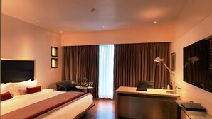 Ein Hotelzimmer mit einem großen Bett, einem Schreibtisch, einem Stuhl, einer Schreibtischlampe, einem Fernseher und einem fenster mit Vorhängen.