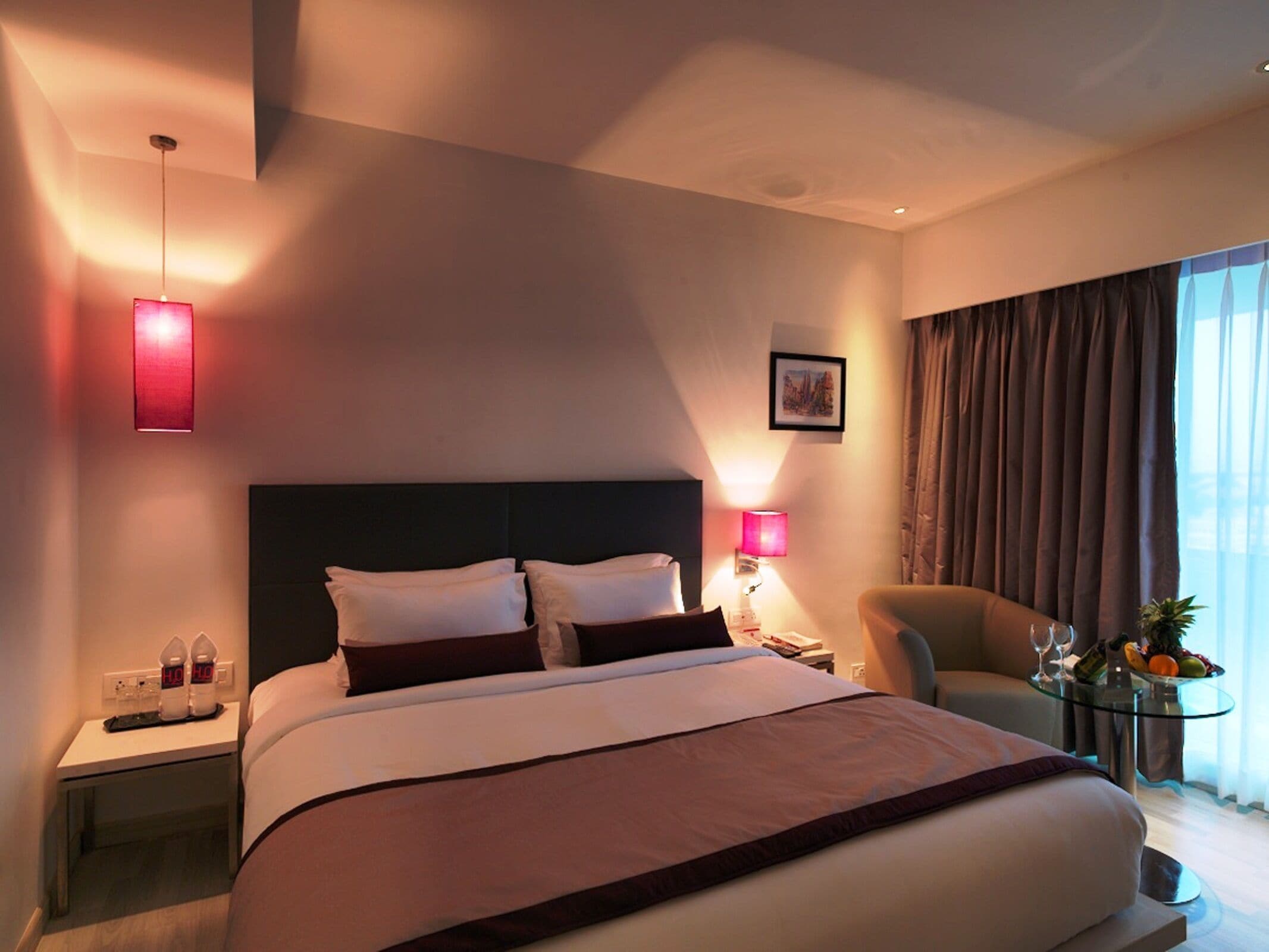 A modern hotel room with a large bed, bedside tables, a chair, a small table with a fruit bowl and glasses, and a window with curtains.