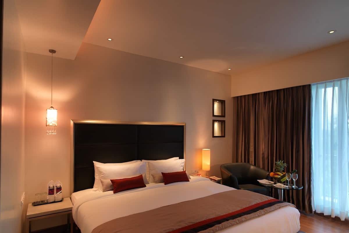 Een hotelkamer met een groot bed, een nachtkastje met een lamp, een stoel, en een klein tafeltje met fruit en glazen.