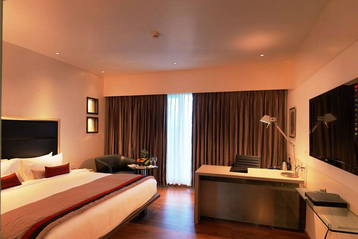 Een hotelkamer met een groot bed, een bureau, een stoel, een klein tafeltje met een fruitmand en een groot raam met gordijnen.