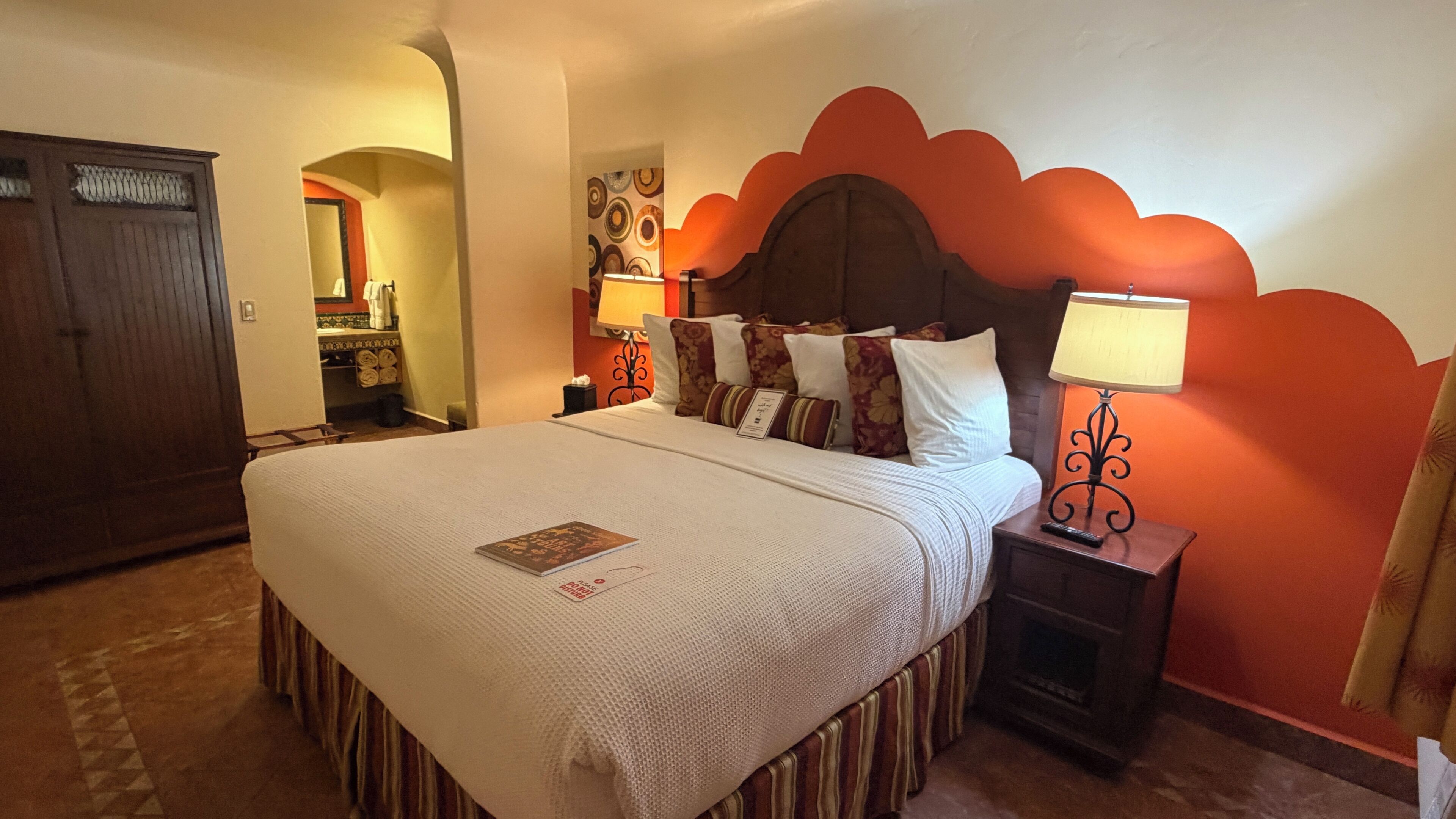 A hotel room with a large bed, a nightstand with a lamp, a wardrobe, and a bathroom visible through an archway.