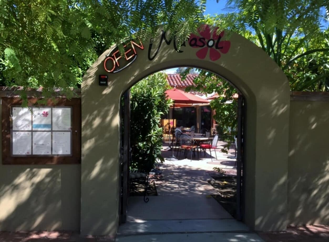 An archway with the words "OPEN" and "Masol" on it, leading to an outdoor seating area with tables and chairs.