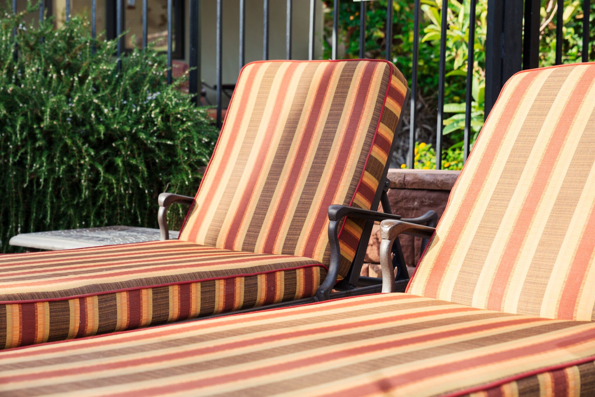 Un conjunto de sillones a rayas con reposabrazos, ubicados al aire libre con una valla metálica y follaje al fondo.