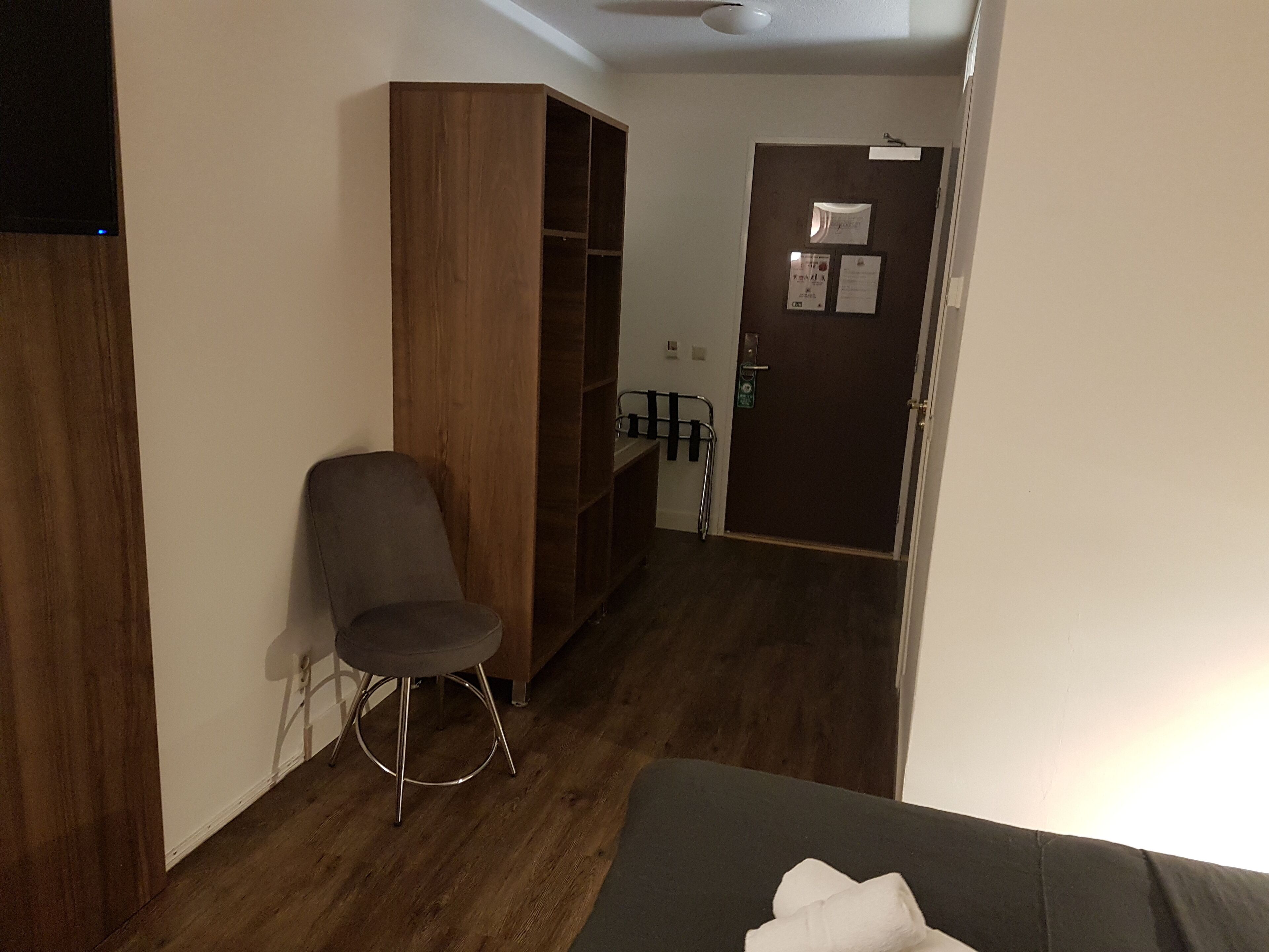 A room with a wooden wardrobe, a grey chair, a small stool, and a bed with a white towel.