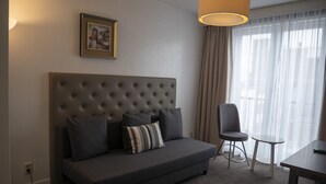 A modern hotel room with a tufted headboard sofa, a small table, a chair, and a large window with sheer curtains. - Hotel Hague Center (The Hague)