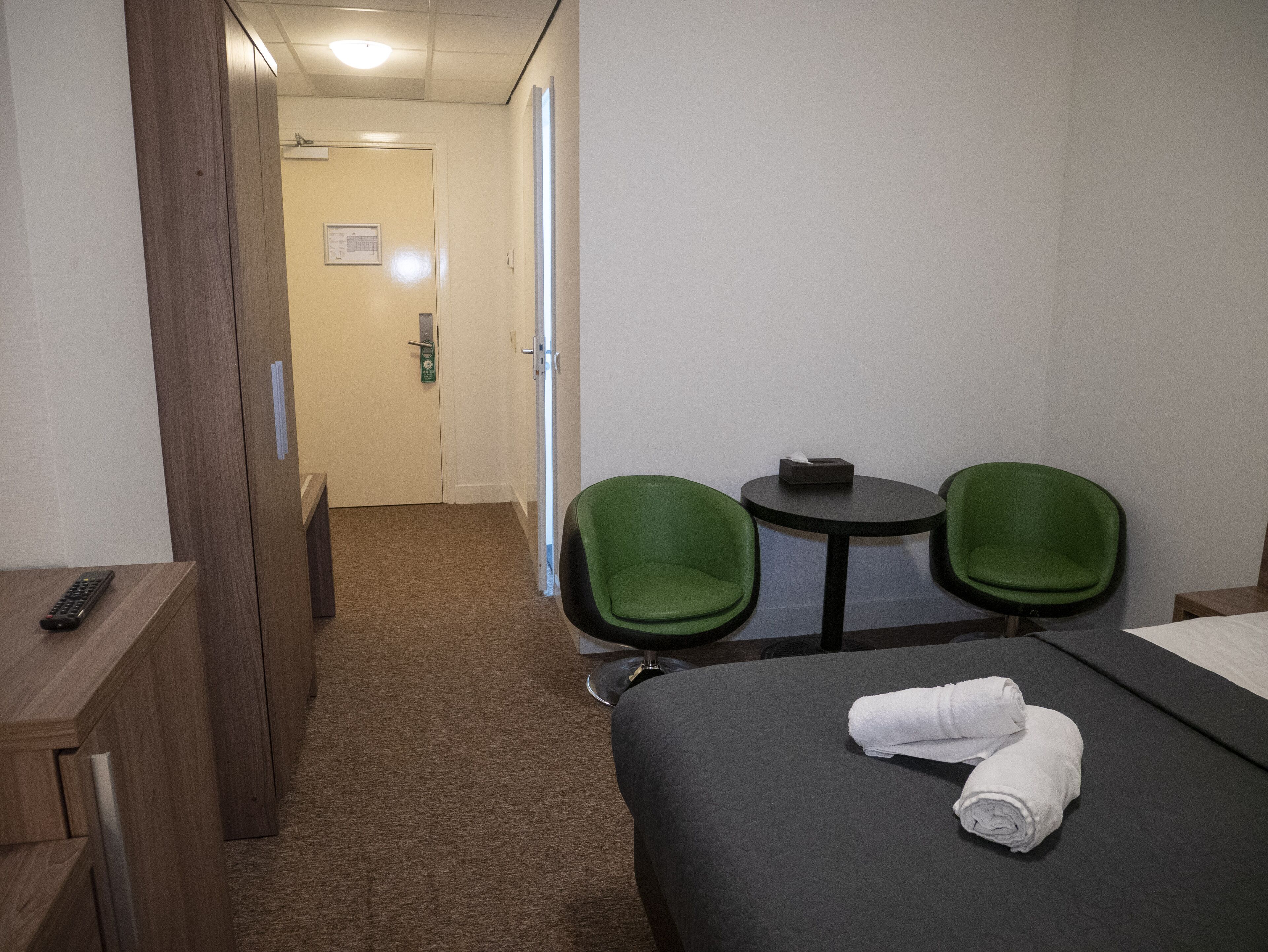 Hotelkamer met een bed, twee groene stoelen, een klein tafeltje en een bureau met een afstandsbediening.