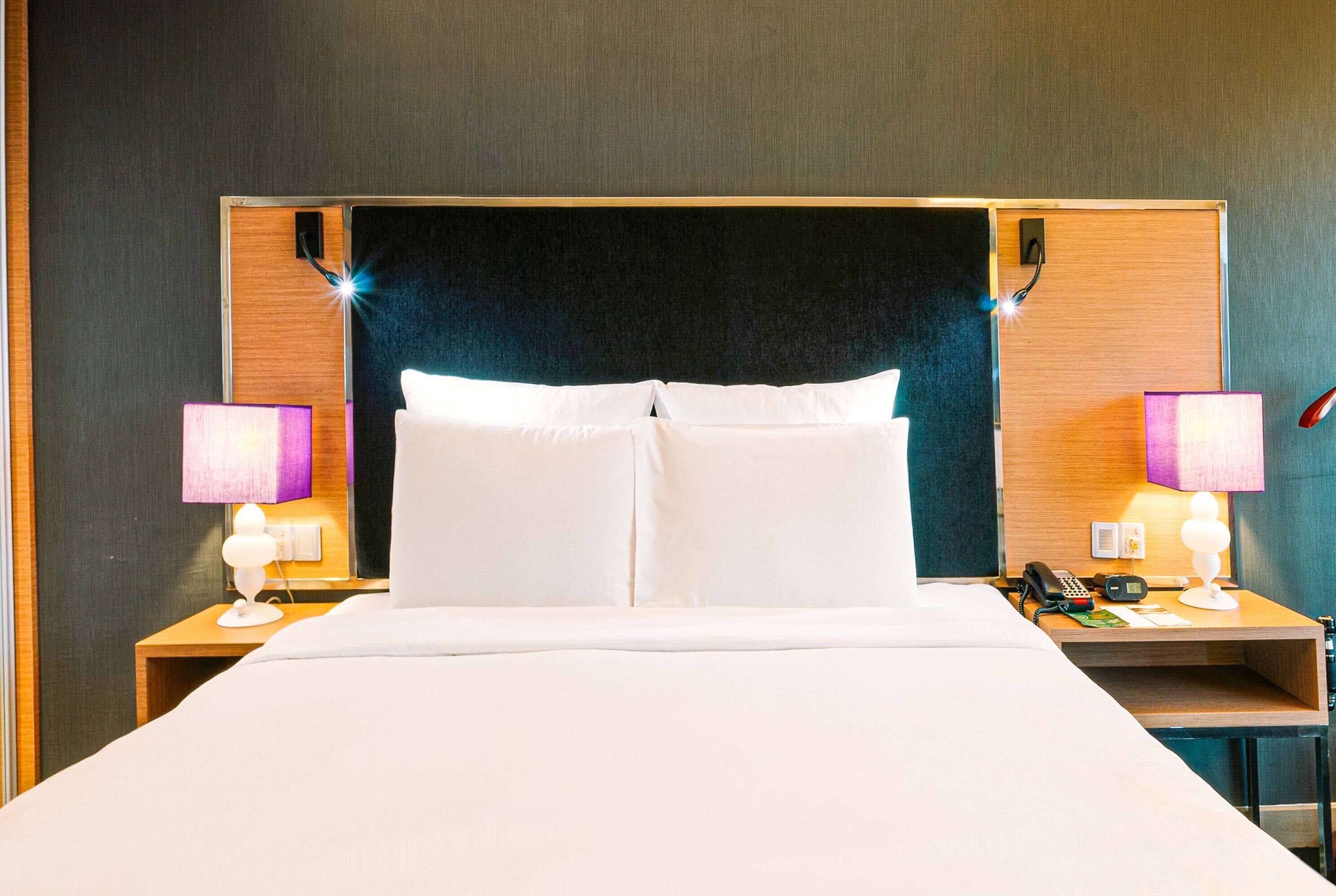 Une chambre d’hôtel avec un grand lit, deux tables de chevet, une petite table, une lampe, un téléphone et un luminaire fixé au mur.