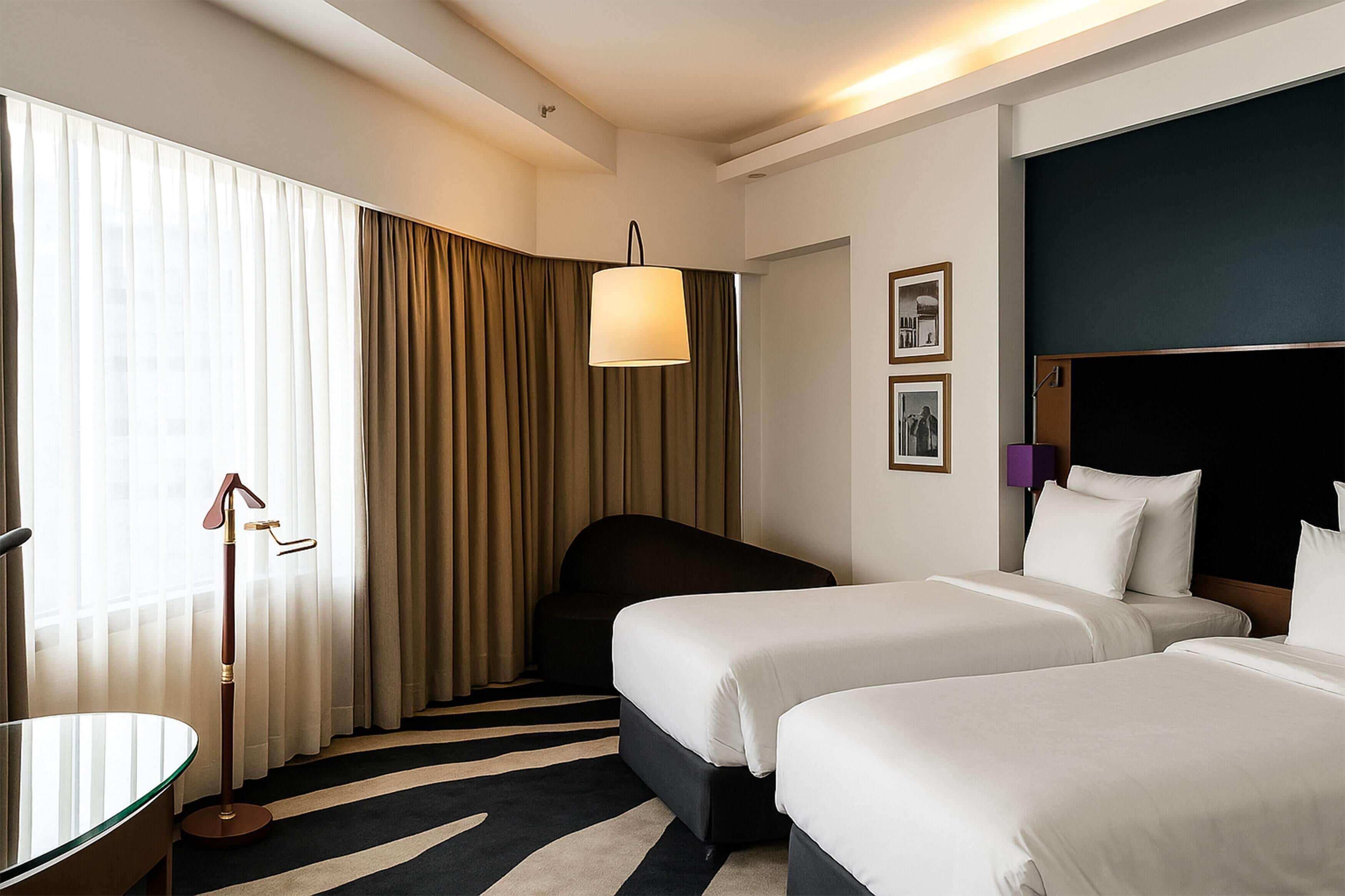 Habitación de hotel con dos camas, un escritorio, una silla y un ventanal grande con cortinas.