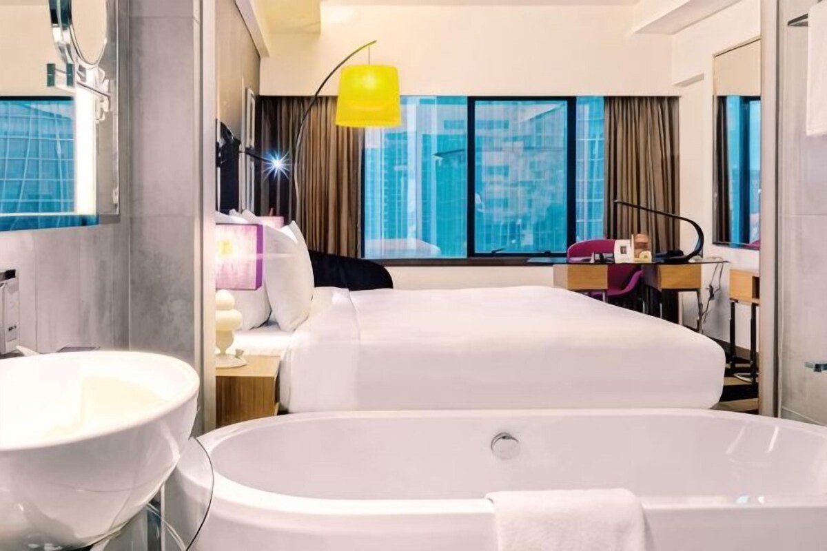 A hotel room with a large bathtub, a double bed, a bedside table with a lamp, a window with curtains, and a city view.