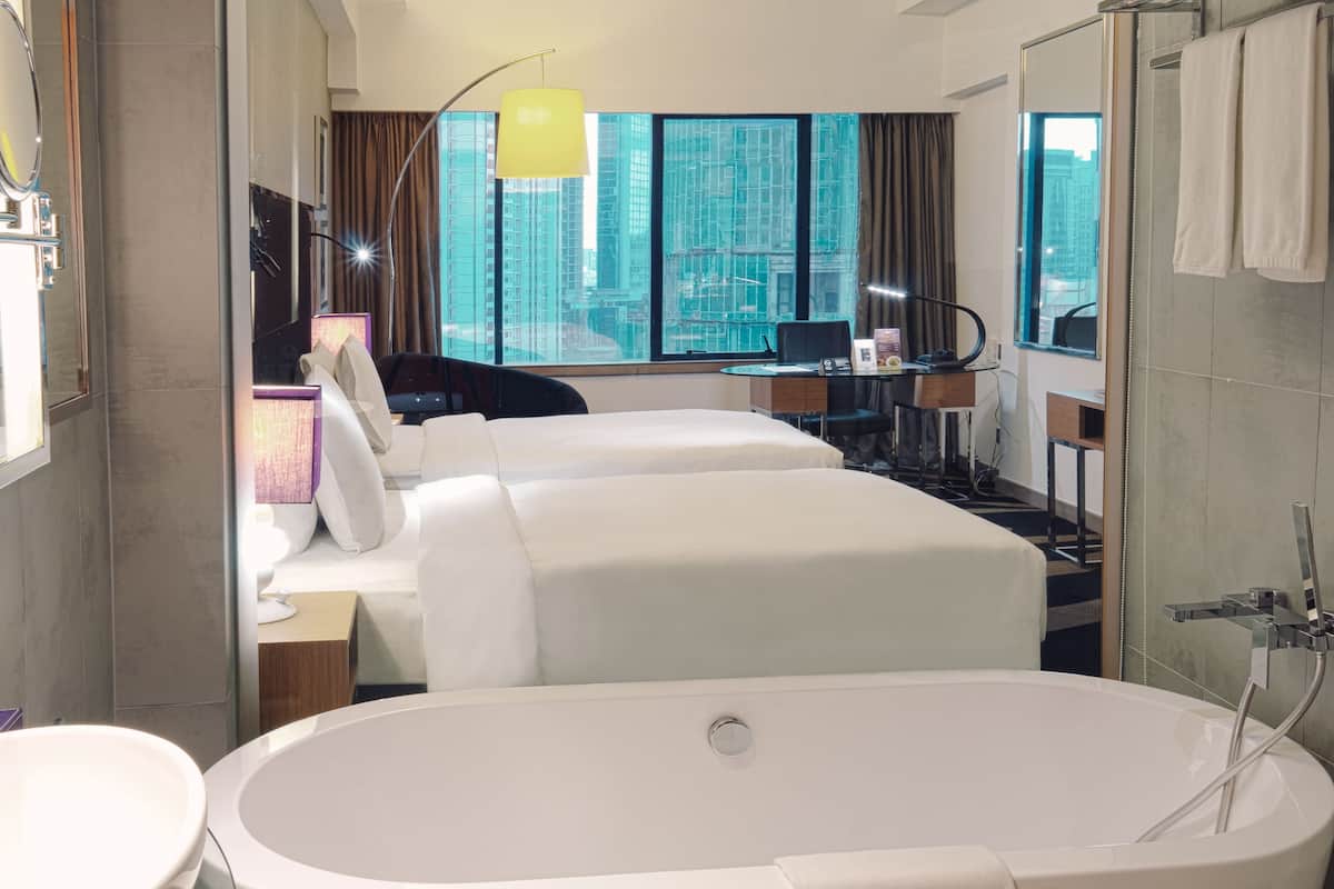 A modern hotel room with a large bathtub, two beds, a desk, and a view of tall buildings through large windows.
