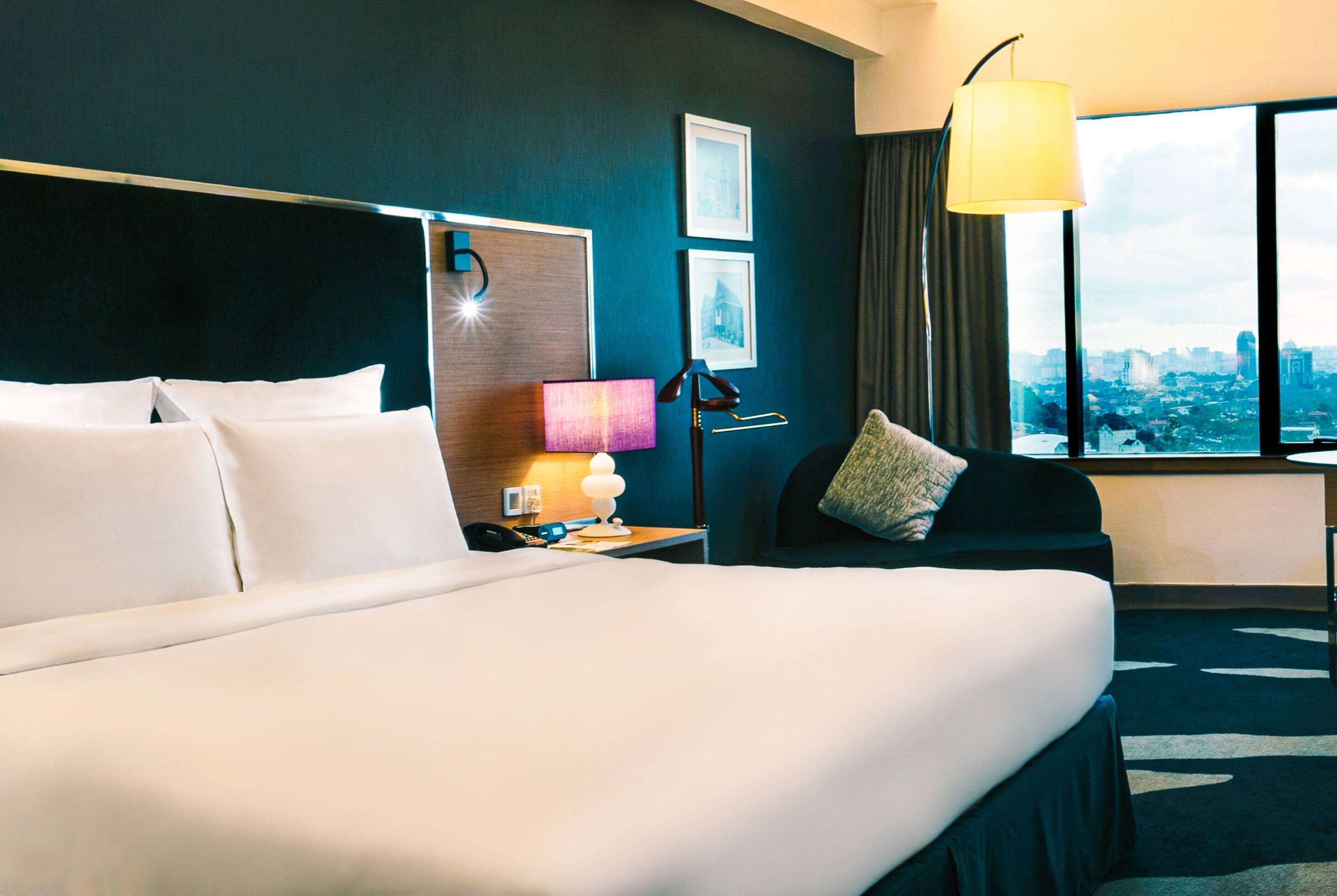 A hotel room with a large bed, a desk with a lamp, a chair, and a view of a cityscape through a window.