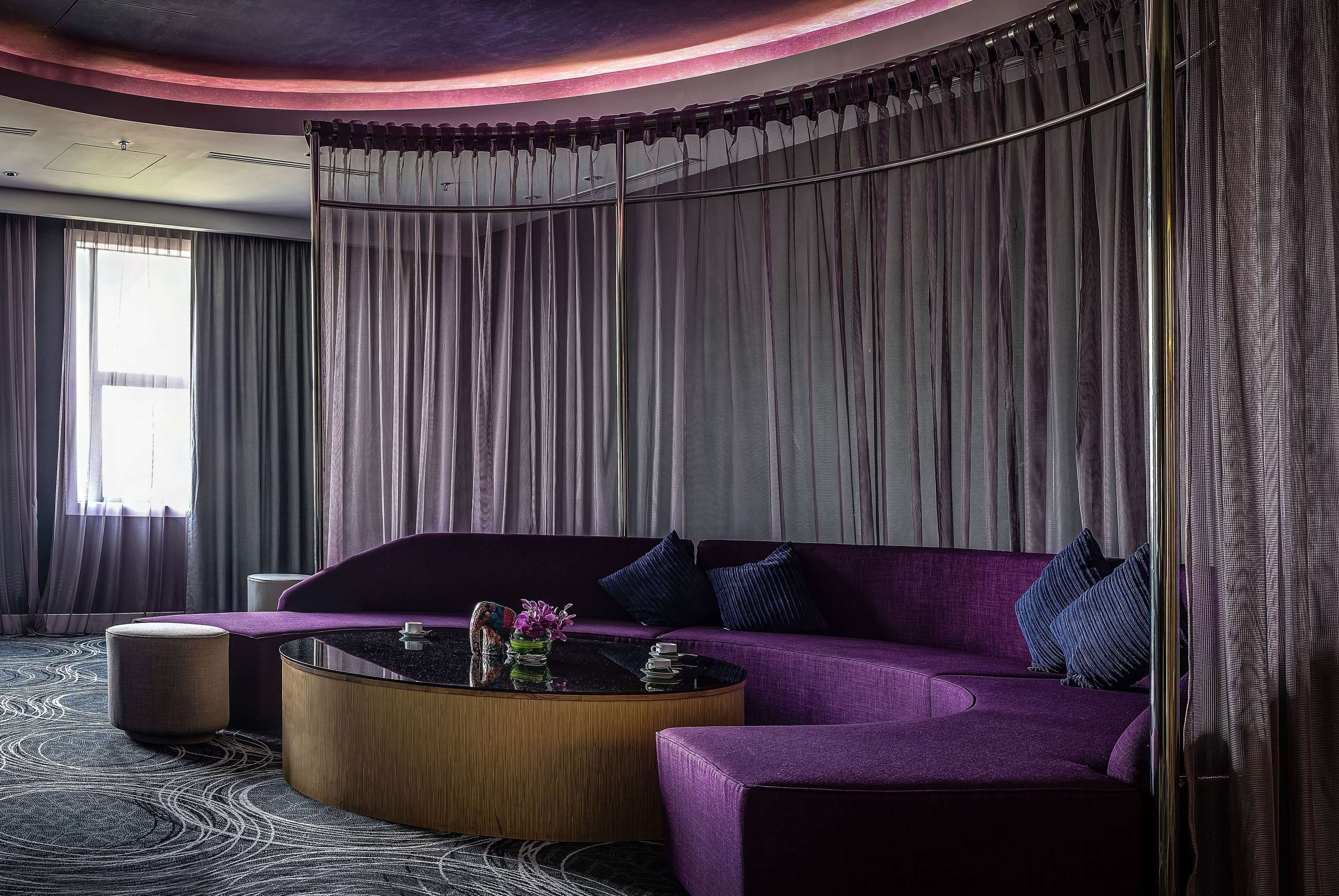 Un salon moderne avec un canapé violet, une table basse ronde et une grande fenêtre avec des rideaux.