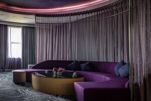 Una sala moderna con un sofá morado, una mesa de centro redonda y un ventanal con cortinas.