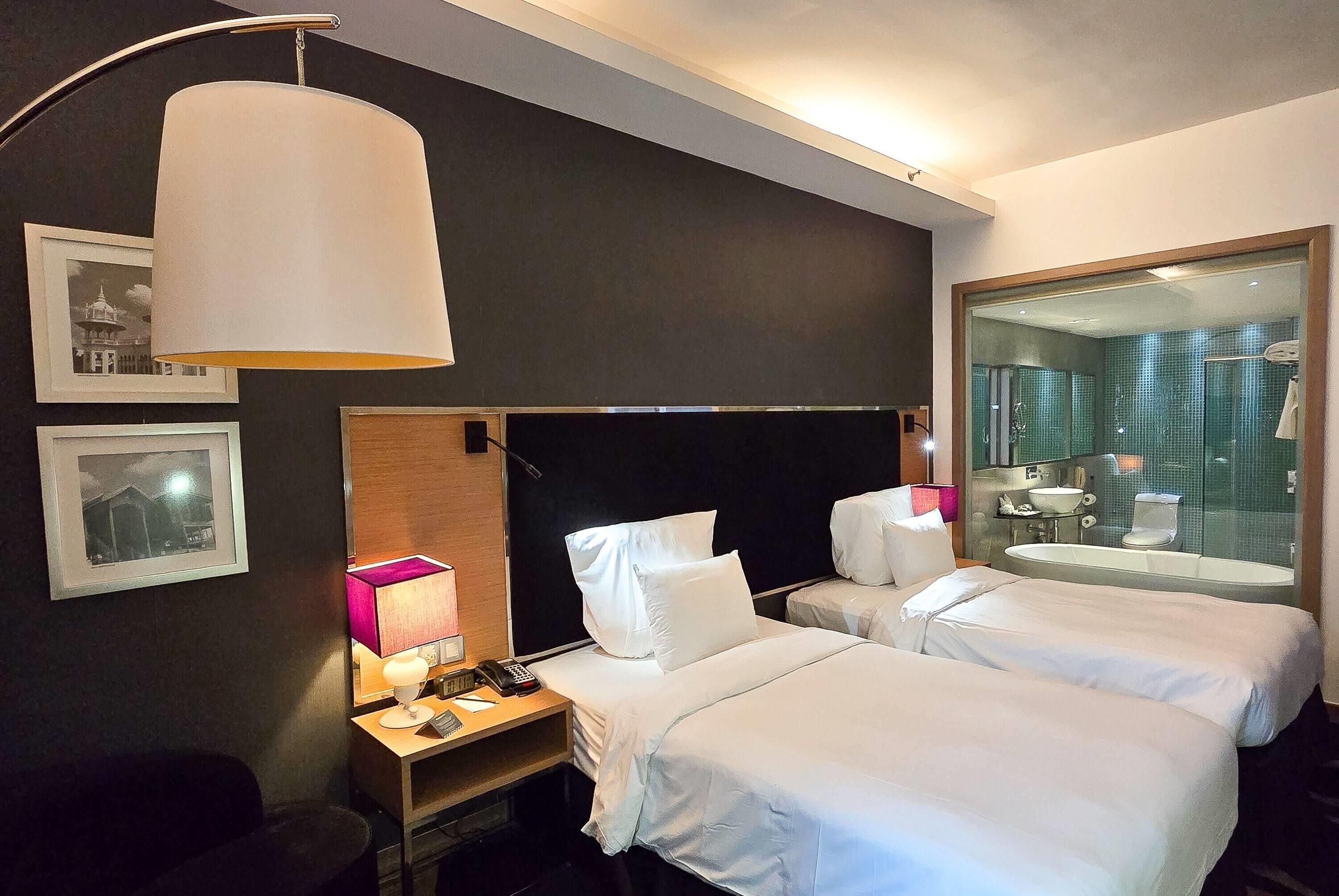Een hotelkamer met twee bedden, een nachtkastje met een lamp, een spiegel en uitzicht op de badkamer.