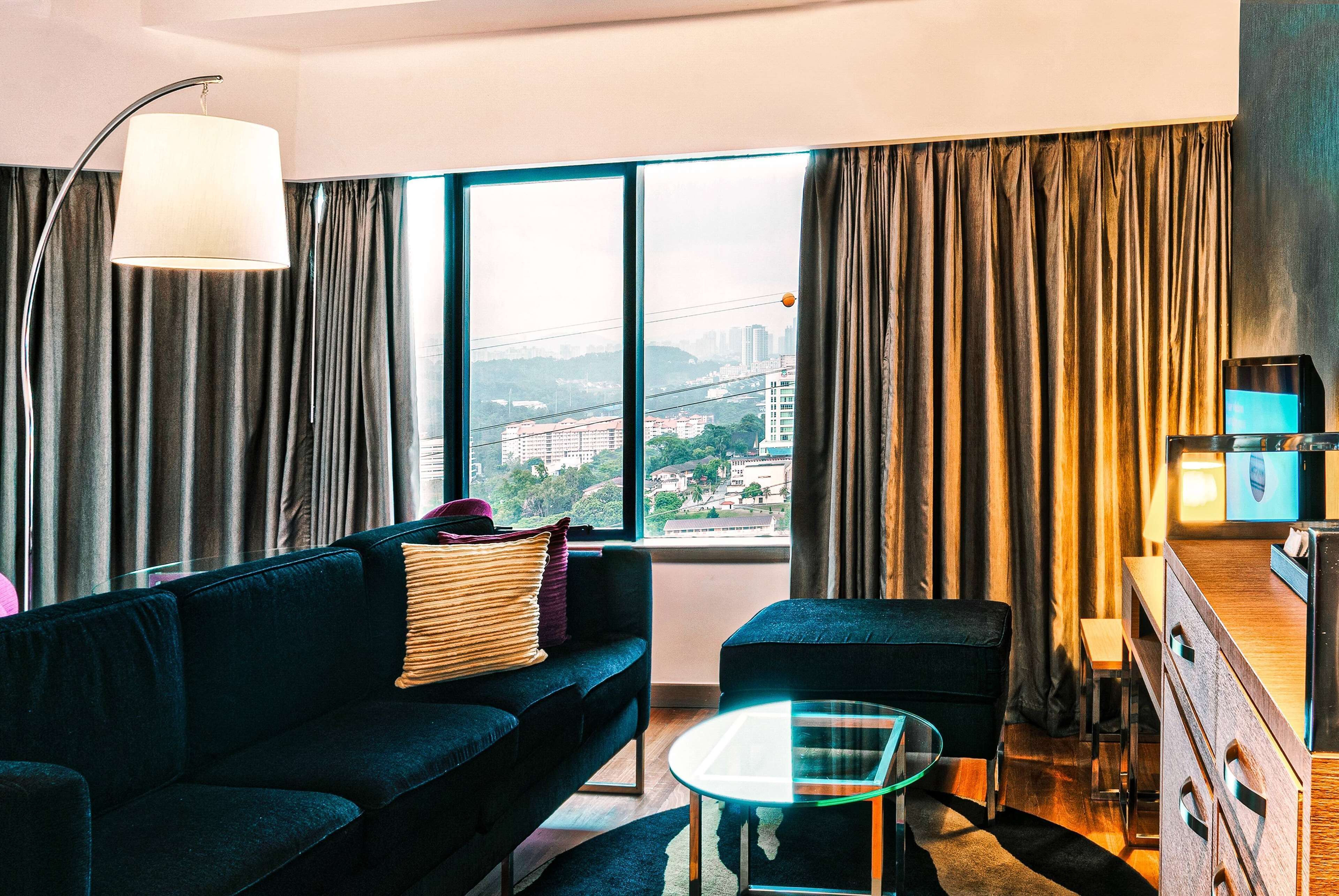 Una sala de estar moderna con un sofá, una mesa de vidrio y vistas a un paisaje urbano a través de amplios ventanales.