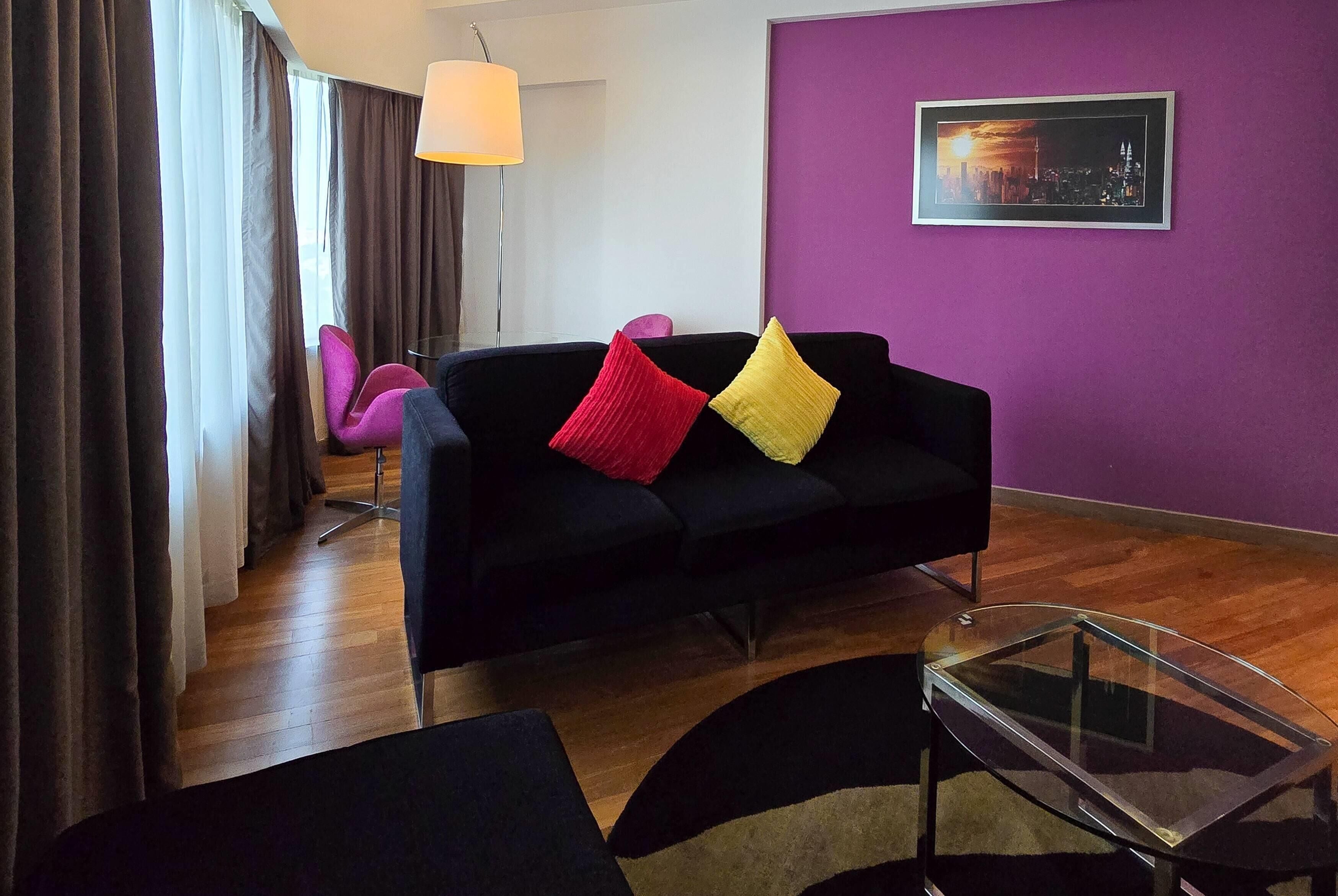 Un salón moderno con un sofá negro, cojines coloridos, una mesa de centro de cristal y una pared morada con una pintura de un paisaje urbano.