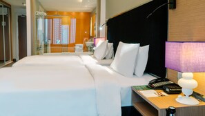 Quarto de hotel com duas camas, uma mesa com telefone e um banheiro visível através de um painel de vidro.