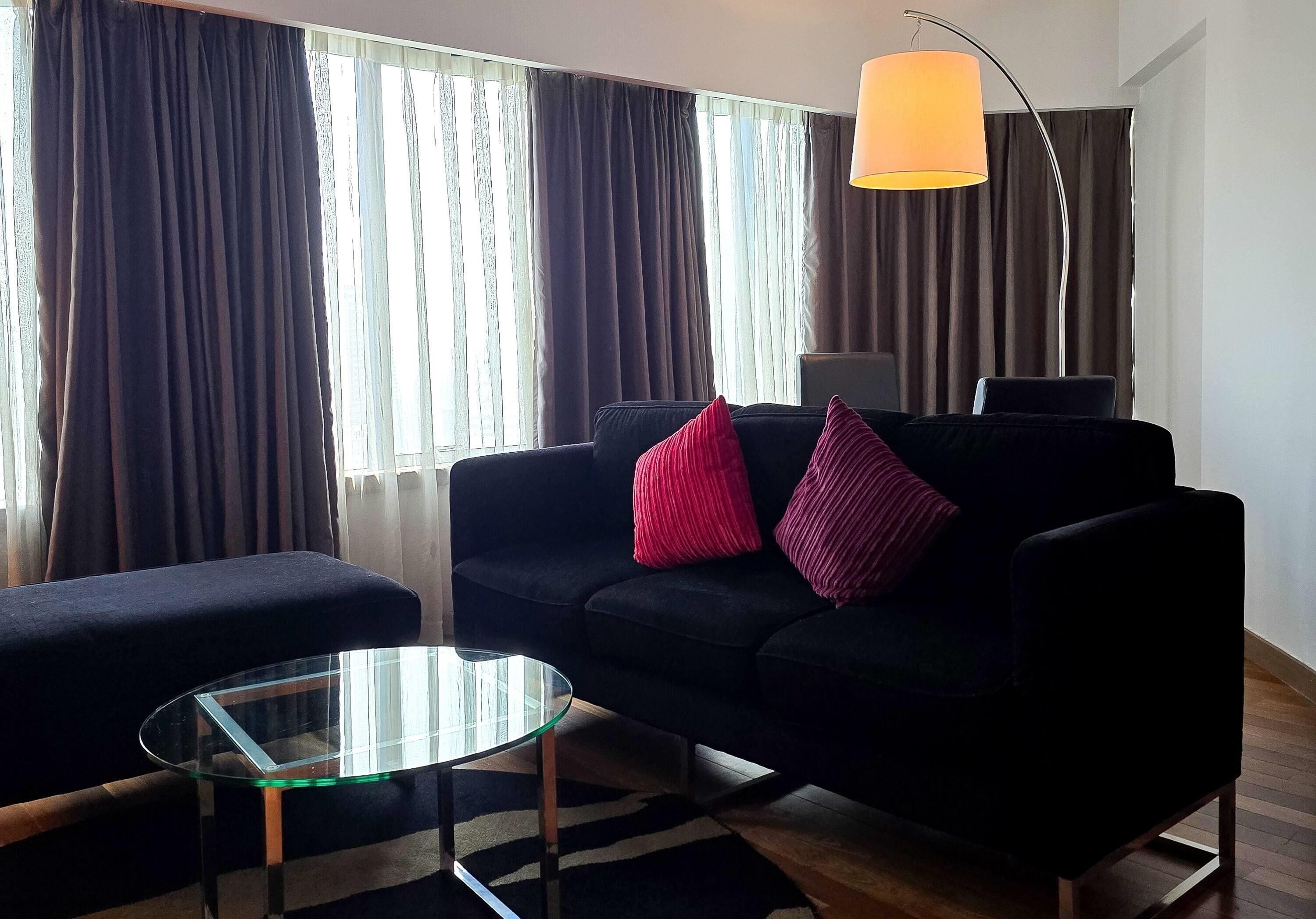 A modern living room with a black sofa, glass coffee table, and a large window with curtains.