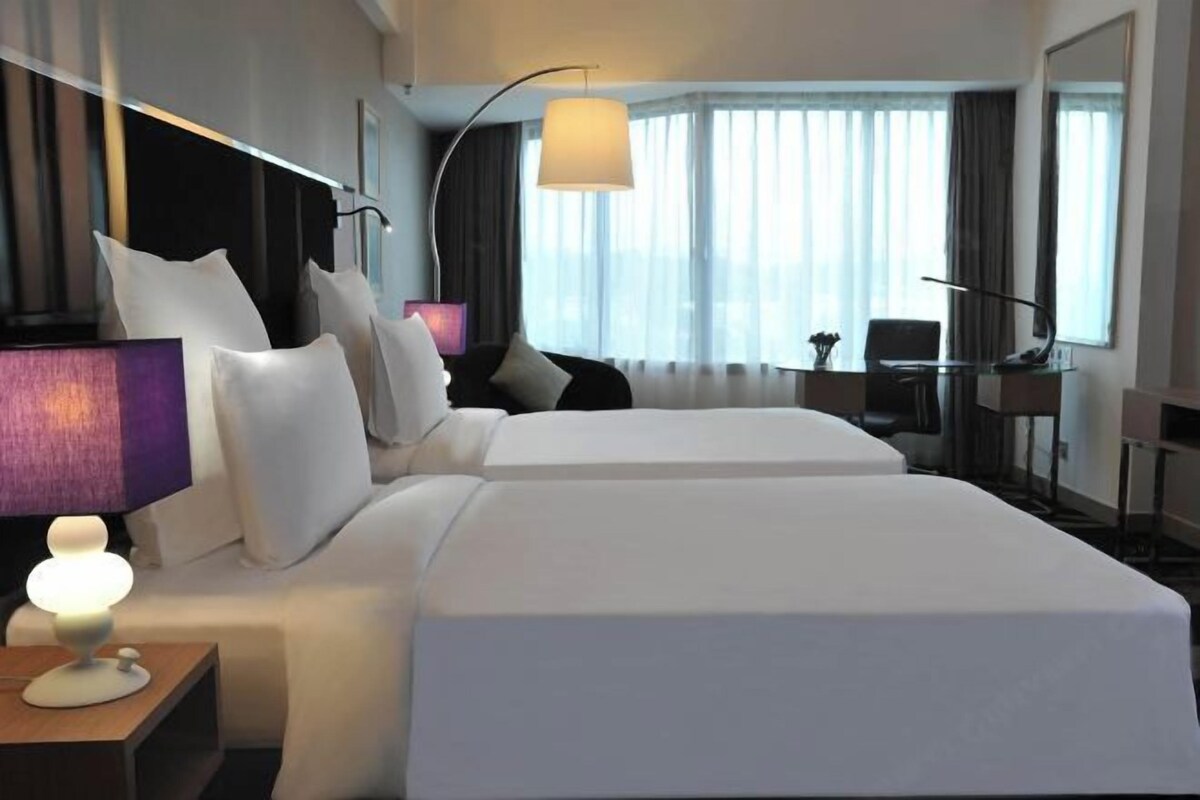 A hotel room with two beds, a desk, a chair, a lamp, and a view of the city through large windows.