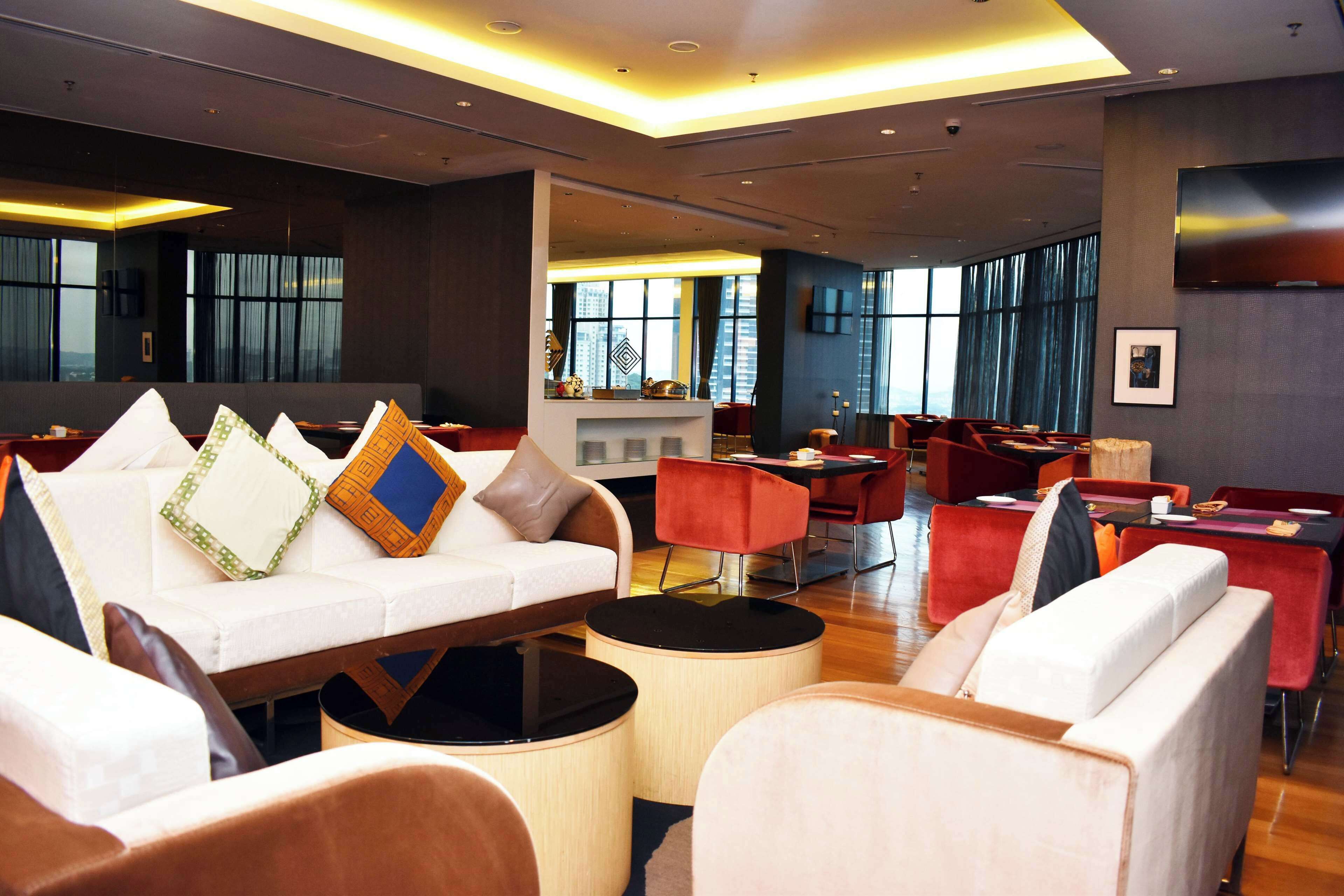 A modern hotel lobby with a sofa, red chairs, a bar area, and large windows.